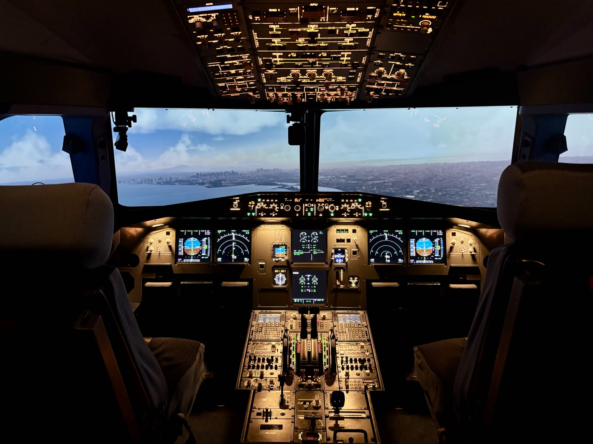 Wide symmetric view of the A320 fixed-base simulator cockpit, dual pilot seats and full glareshield and throttle console, with a sweeping coastal city and bay skyline on the visual displays outside the windscreen.