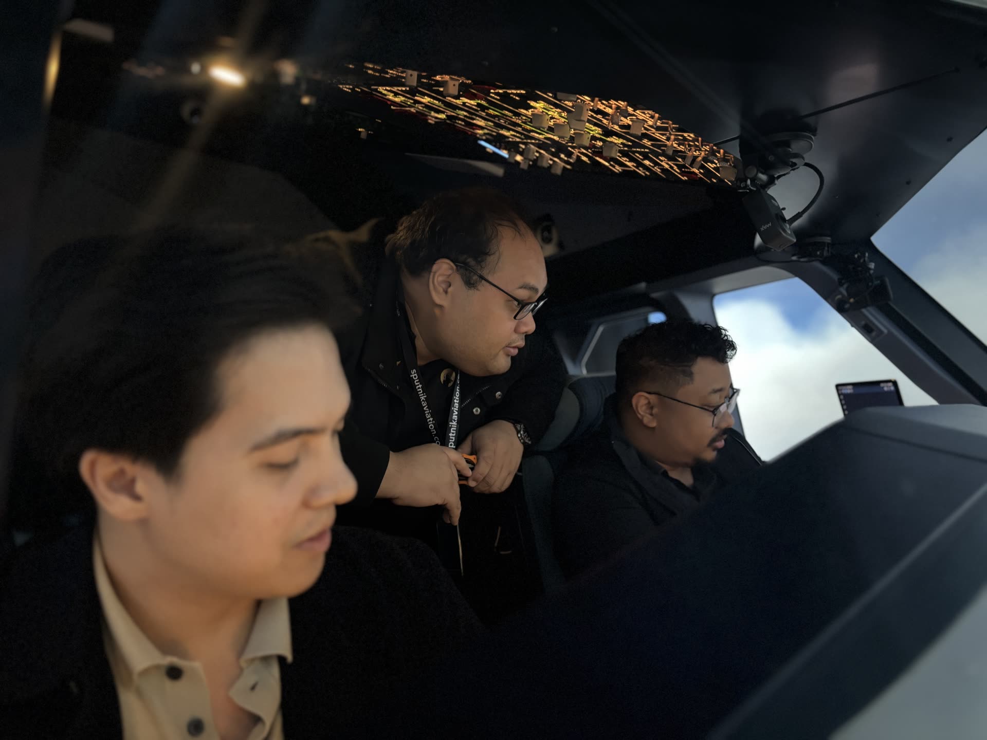 Three Sputnik Aviation guests and staff seated in the A320 simulator flight deck at dusk, the lit overhead panel glowing above them and a Sputnik Aviation lanyard visible on one guest.