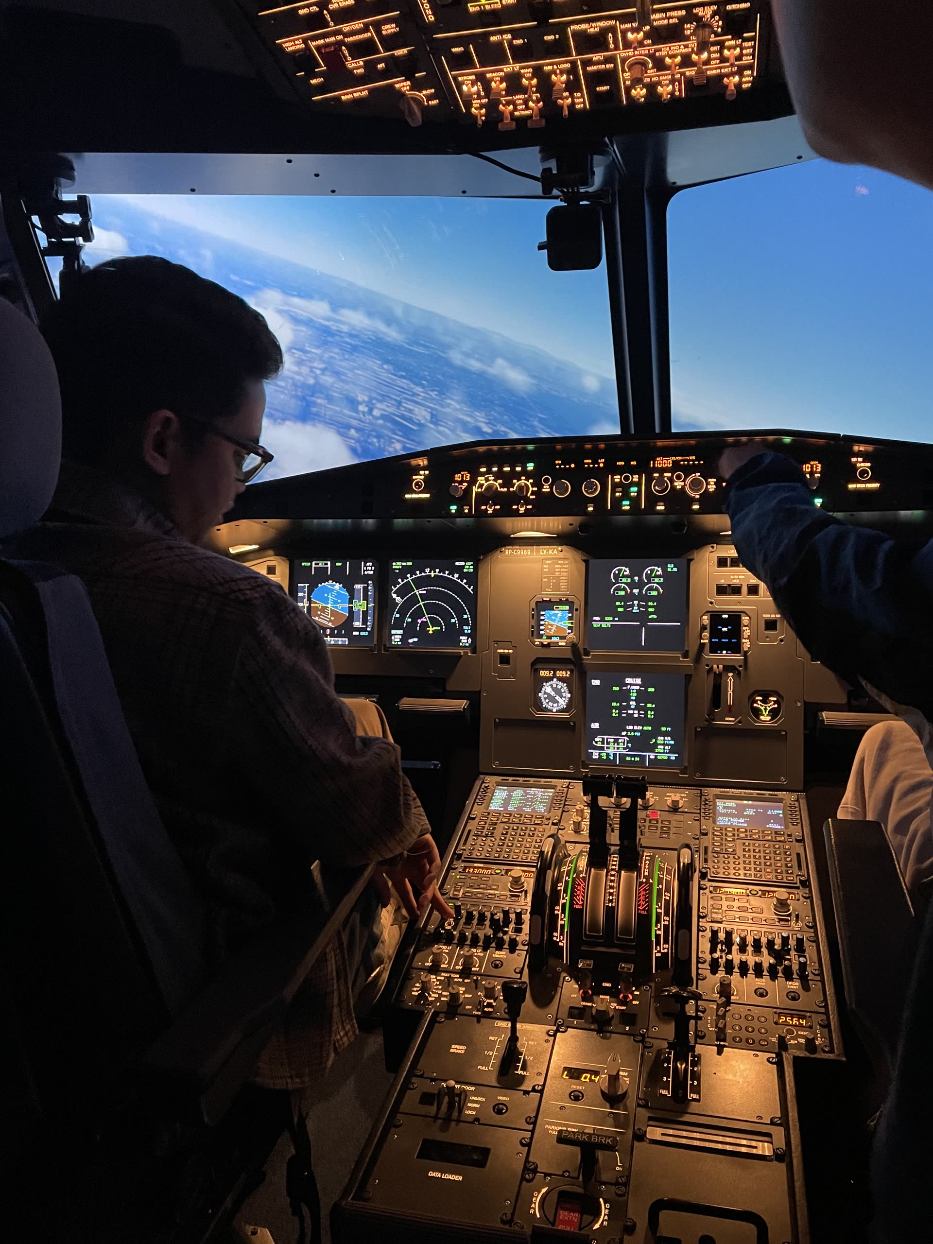 A pilot seated at the A320 simulator controls at dusk, reaching for the flight control unit with the overhead panel lit and a warm sunset sky filling the outside visual.