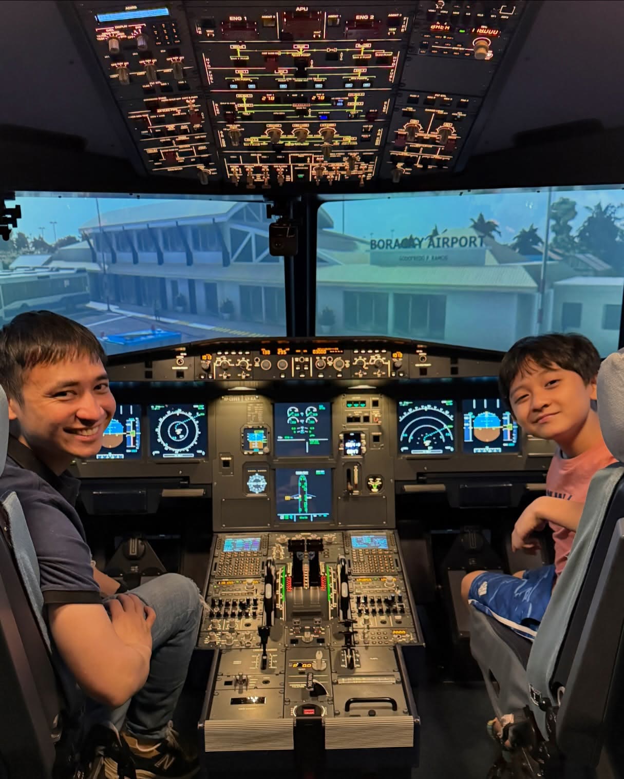 A father and son seated together at the A320 flight deck simulator controls, smiling back at the camera with Boracay Airport shown on the outside visual.