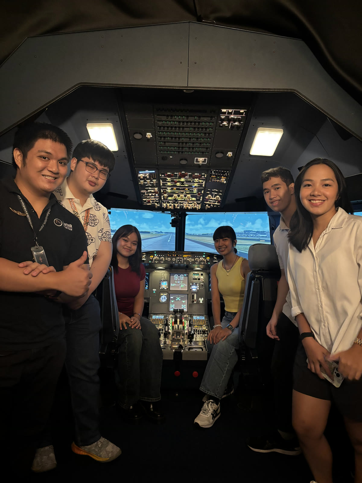 A group of six friends and staff gathered around the A320 flight deck simulator controls, smiling together with the runway visual lit up outside.